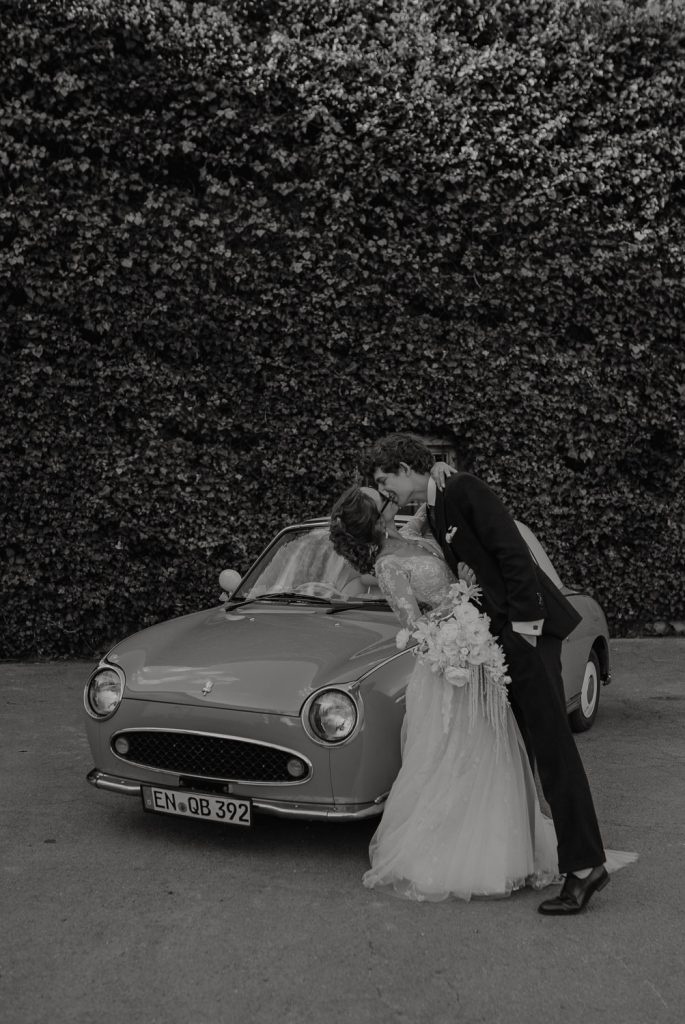 Brautpaar küsst sich vor dem Oldtimer ihrem Brautaut in Schwarz weiß