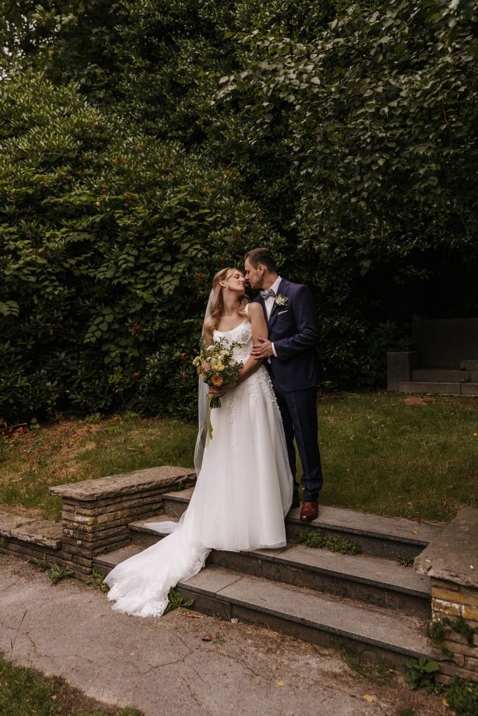 Brautpaar steht auf einer Treppe im Botnaischen Garten. Braut hält einen Brautstrauß in den Händen und lehnt sich an ihren Bräutigam