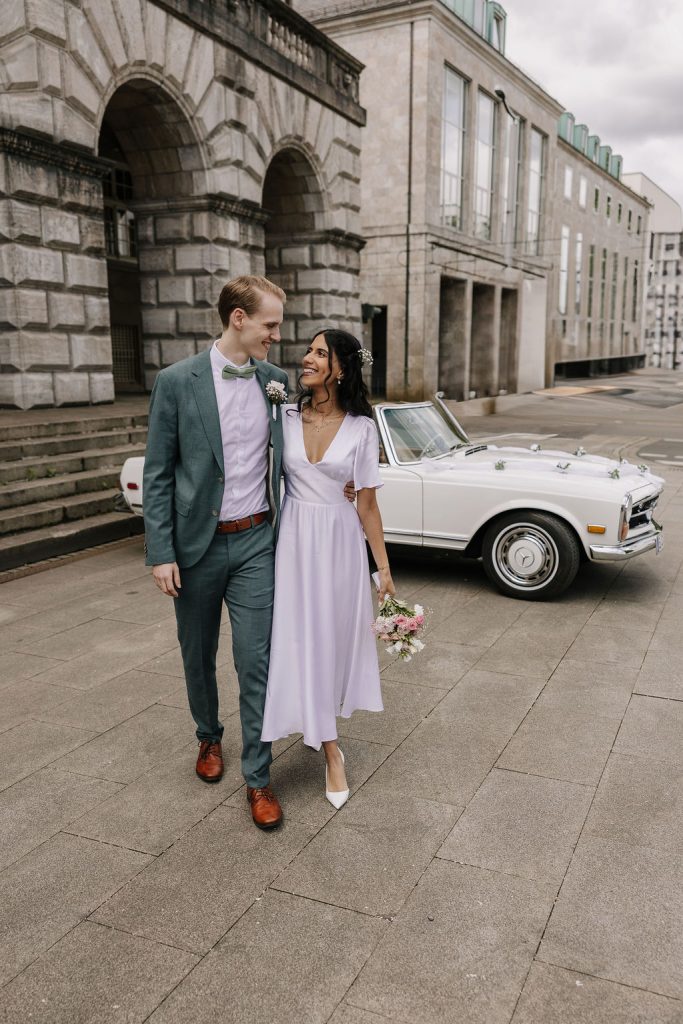 Brautpaar läuft im Anzug und Brautkleid mit einem Brautstrauß an ihrem Hochzeitstag entlang. Hinter ihnen steht das Brautauto