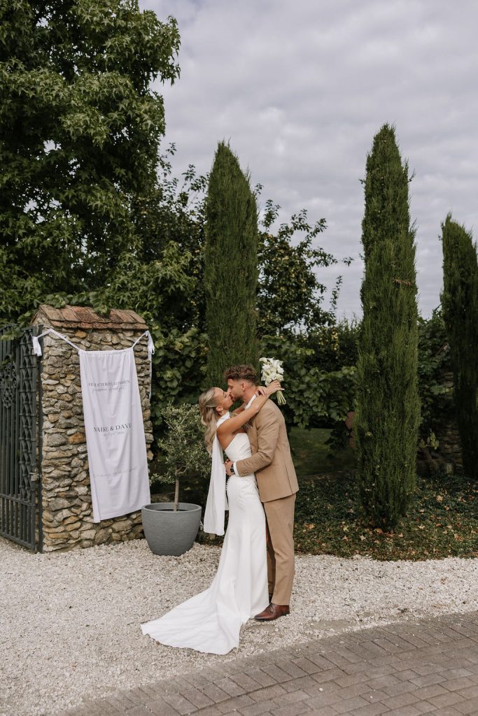 Brautpaar küsst sich vor einem Steintor im grünen Garten des Kloster Kraul, romantisches Paarshooting nach der Hochzeit im Freien.