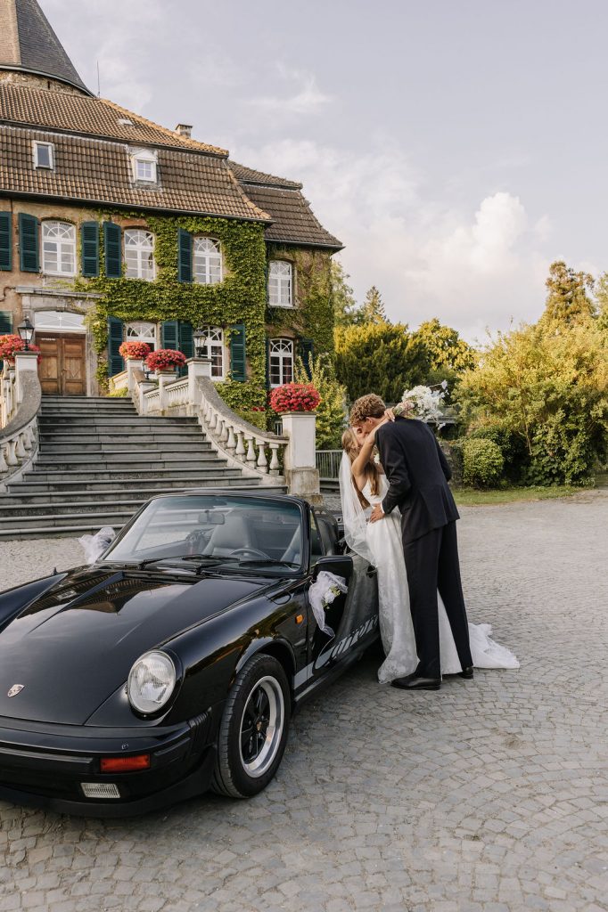 Moderne und elegante Hochzeit in Ratingen – Brautpaar beim Paarshooting vor Schloss Linnep mit klassischer Architektur, Natur und zeitlosem Editorial Look. Snd angeekelt an Hochzeitsauto Porsche